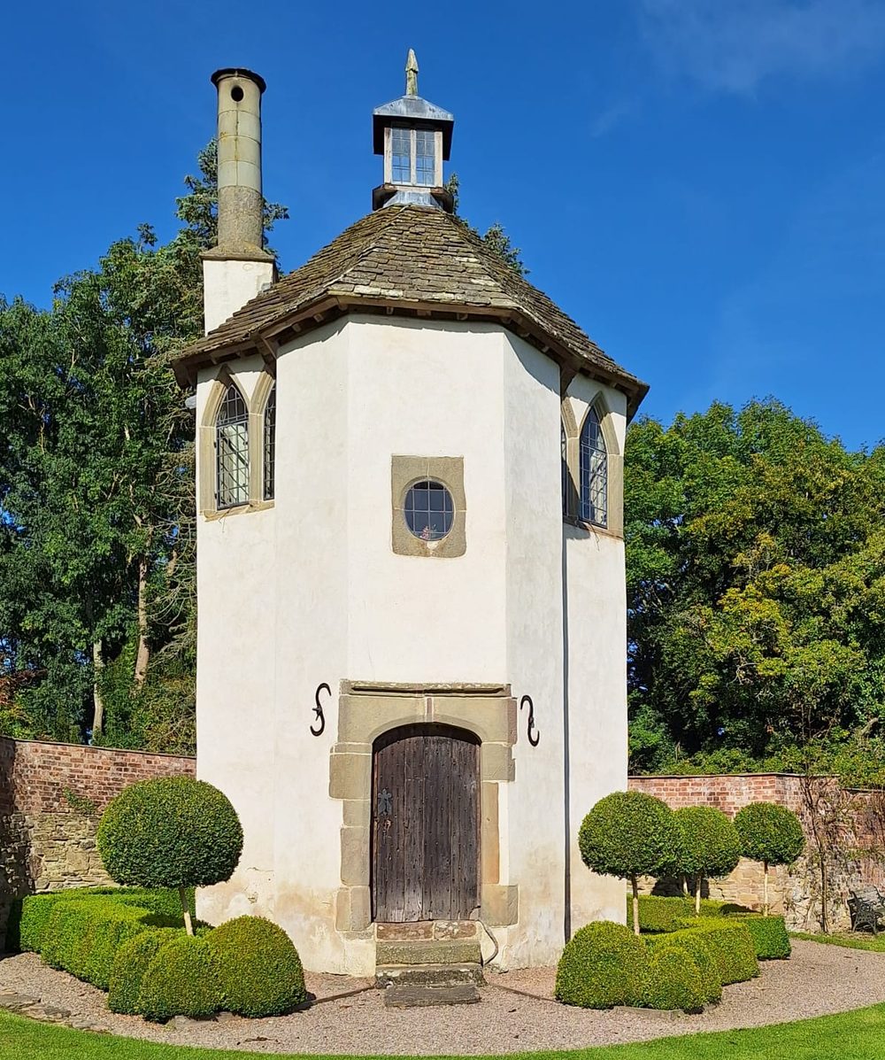 Octagonal white garden house with a shingled roof and cupola among clipped topiary