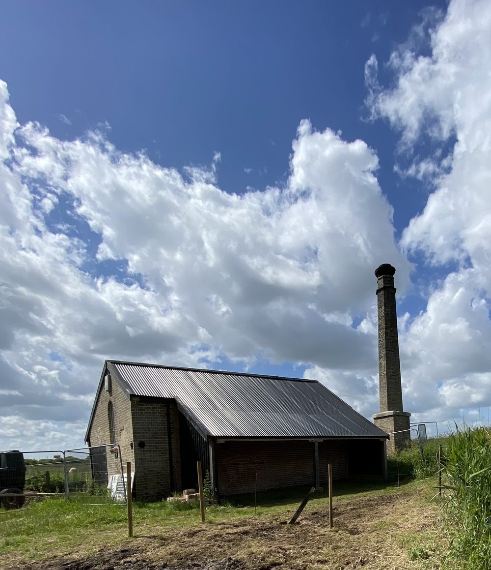 Old brick steam pumphouse with a corrugated roof and a tall chimney against summer cloud