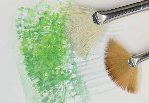 Two fan brushes with different bristle colours, shown next to a textured green swatch created with watercolour pencils, demonstrating their use for foliage or grass effects