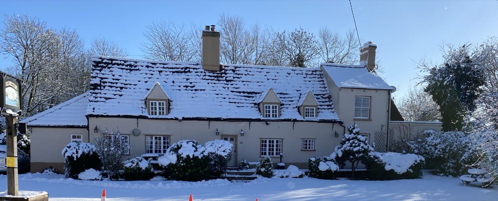Snow-covered tiled roof of a village pub in shadow, the snow reading as cool grey-blue rather than white, with two brick chimneys against a bright winter sky