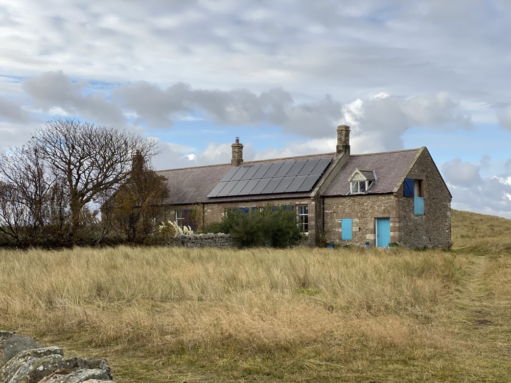 Stone house on the island of Lindisfarne with a block of solar panels covering much of one roof slope and a chimney at each end