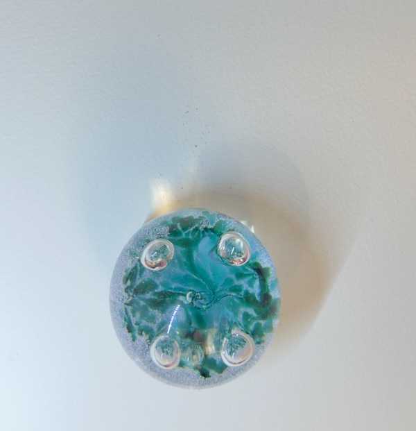 Blue-green glass paperweight under cool light casting faint blue shadow above and subtle orange/warm-toned shadow to the right.