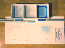 Water added to the shavings (in the palette wells) sitting behind a piece of paper with the relevant swatch below each well. The three pencils are also laying on the paper.