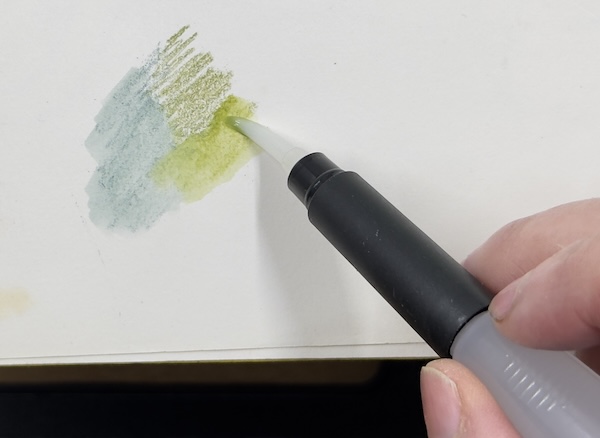 Hand holding a waterbrush, activating green and blue watercolour pencil strokes on paper, showing the pigment dissolving and blending into a wet wash