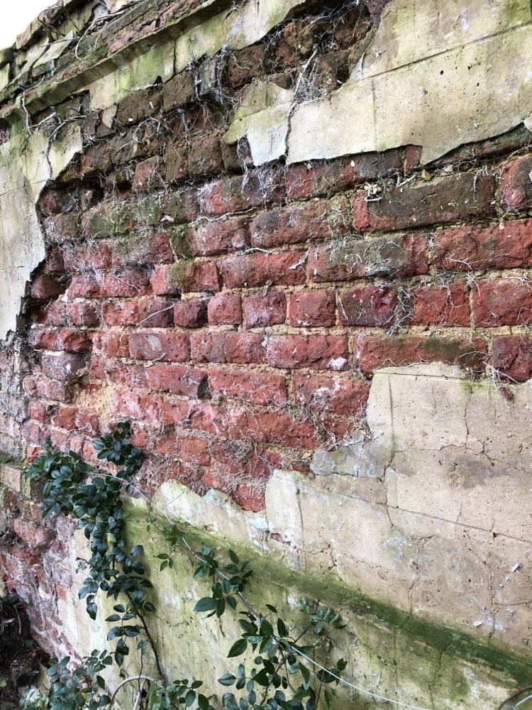 Old wall with render fallen away, exposing broken weathered red brick beneath