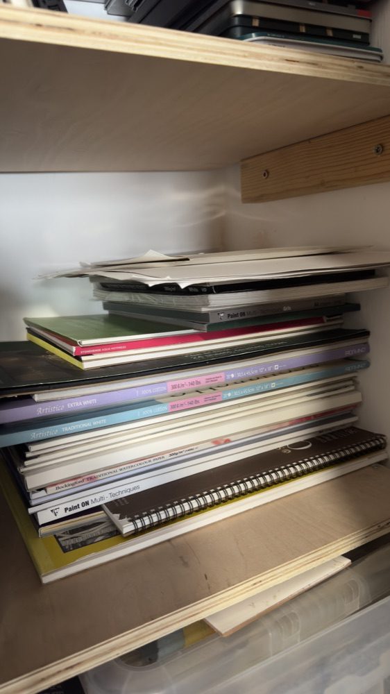 Tall stack of large drawing and watercolour paper pads on a cupboard shelf